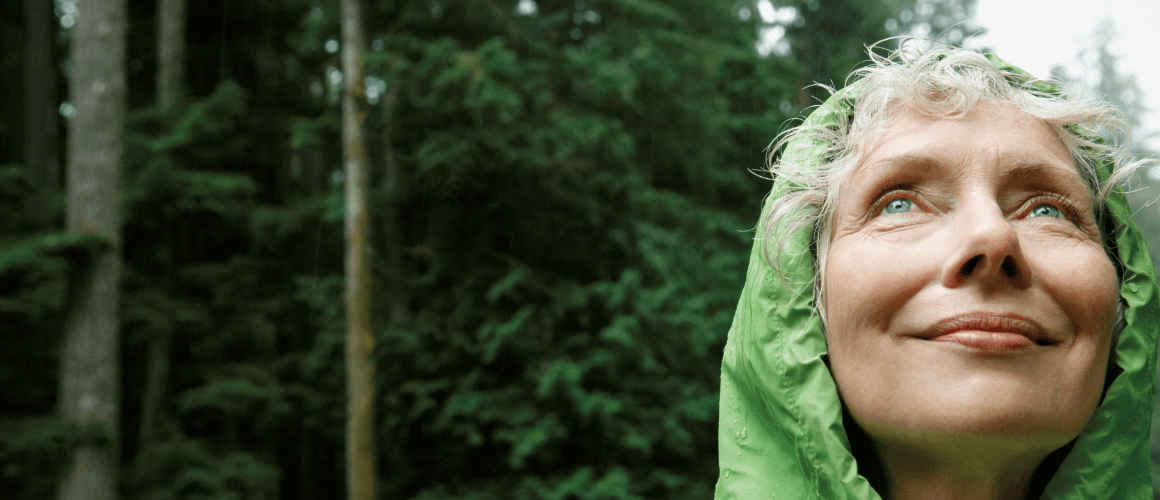 Migraine_Preventive_1160x500.PNG Woman wearing a green rain jacket, standing in a forest and looking up at the sky.