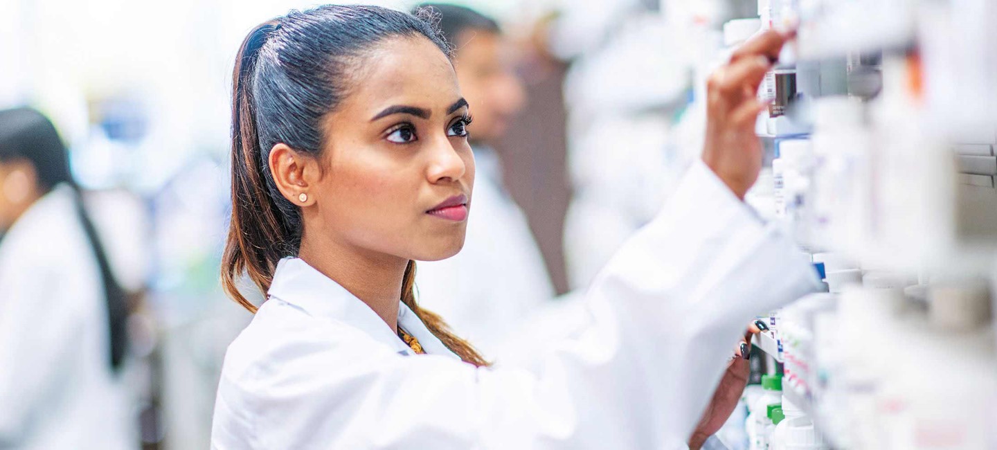 prefforgepants2320x1000.jpg Woman in white lab coat reaching for a bottle of pills on a shelf
